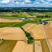 High-angle aerial shot of a patchwork of agricultural fields in a rural landscape. The fields vary in color from light tan to green, suggesting different crops or stages of growth. Scattered farmhouses and outbuildings are visible amongst the fields, connected by dirt roads. Patches of trees and a winding stream add texture to the scene. The horizon shows more fields and distant trees under a blue sky with scattered white clouds.