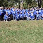 A large group of people wearing matching blue shirts with Day in the Parks printed on them pose together outdoors on a grassy area with trees and tents in the background, under sunny weather.