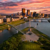 Aerial view of the Dayton, Ohio skyline at sunset, with the Great Miami River and a pedestrian bridge in the foreground and a dramatic orange and pink sky above.