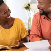 A smiling couple sits at a table in a well-lit home setting, reviewing financial documents together. The woman, wearing a yellow top, holds a pen and a smartphone, while the man, in an orange shirt, holds a piece of paper. A laptop and notebook are visible on the table, creating a collaborative and focused atmosphere. The background shows blurred home decor, adding warmth to the scene.