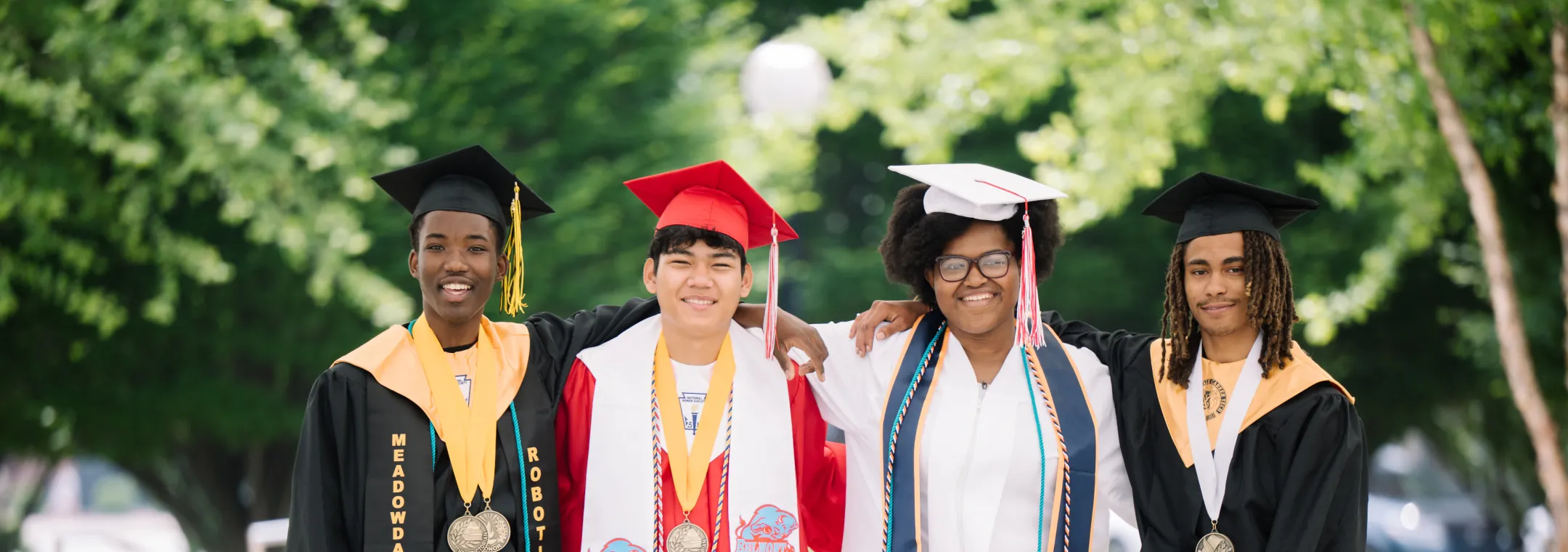 Four diverse high school graduates wearing caps and gowns stand together outdoors, smiling and posing with arms around each other. Each student wears medals and stoles, representing different academic achievements and school spirit.