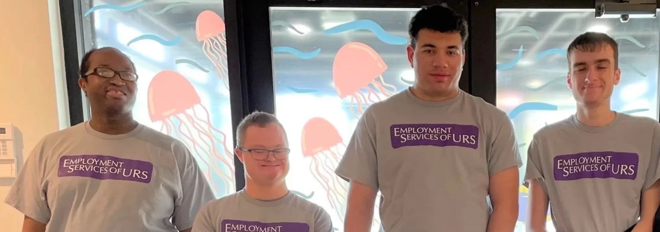 Four people wearing matching gray 'Employment Services of URS' t-shirts stand in a row and smile in front of a window decorated with pink jellyfish and blue waves.