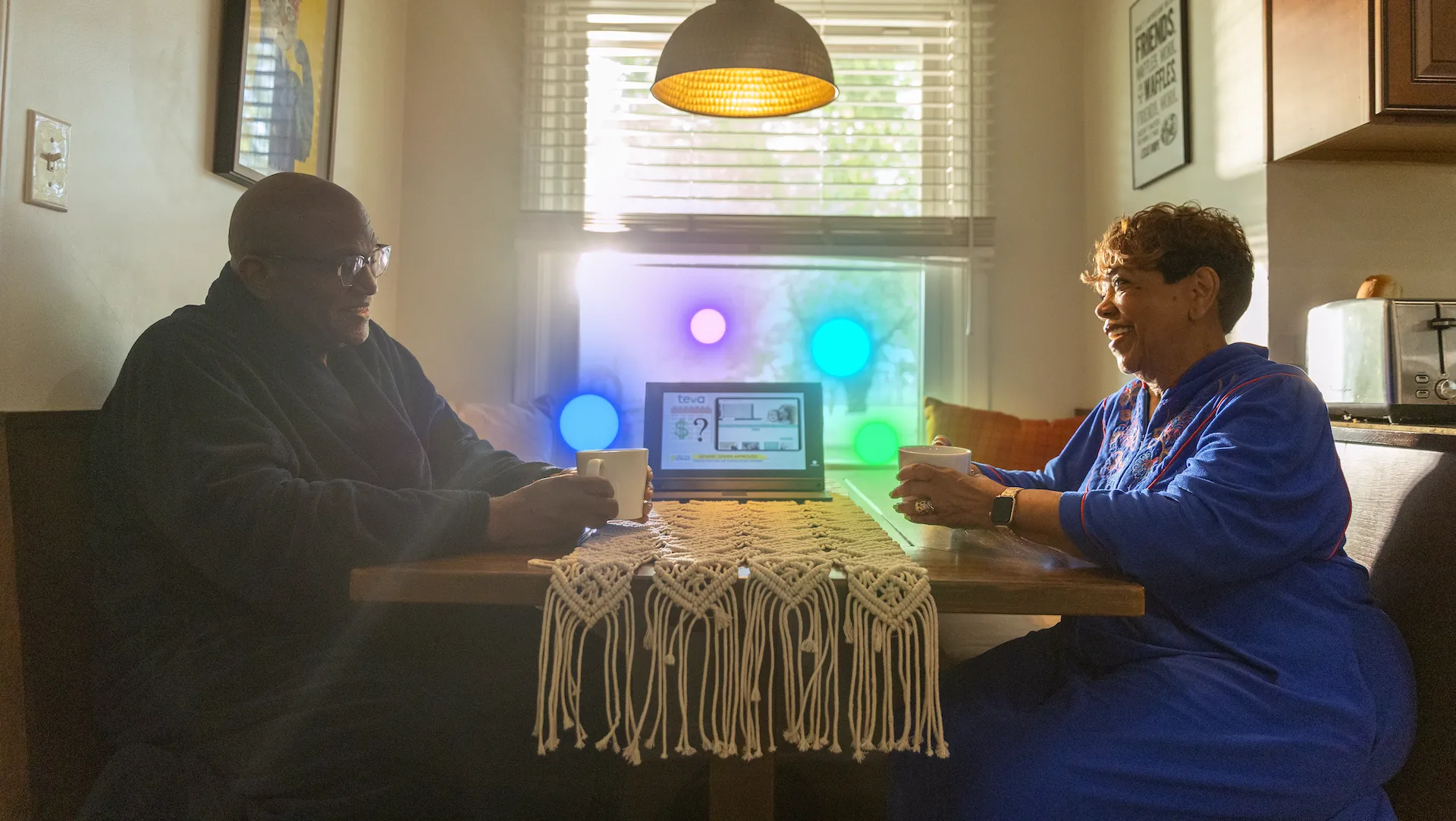 Two people sitting at a wooden kitchen table with a laptop, coffee mugs in front of them, and a macramé table runner. 