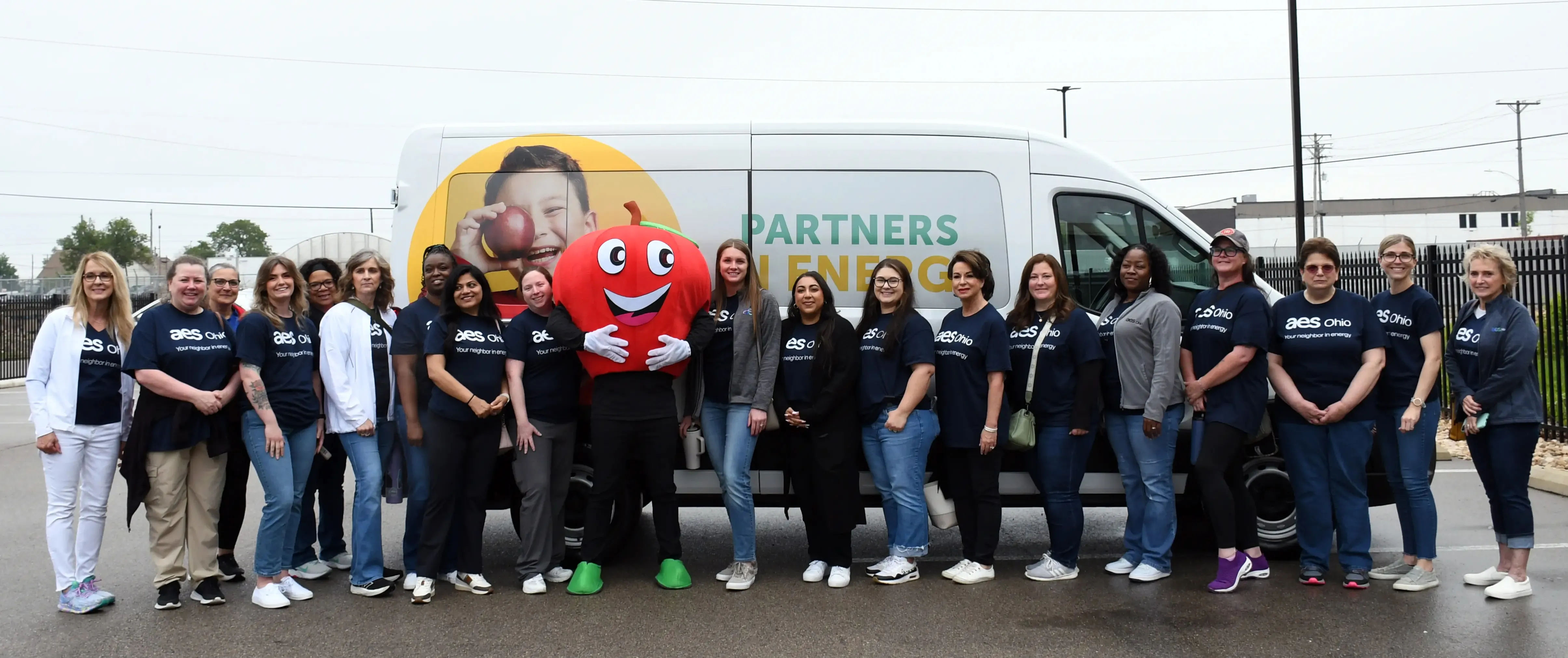A group of people wearing AES Ohio shirts stand in front of a white van with a smiling apple mascot. The van features an image of a child holding an apple and the words 'Partners in Energy.' The group is outdoors on a cloudy day in a parking lot.