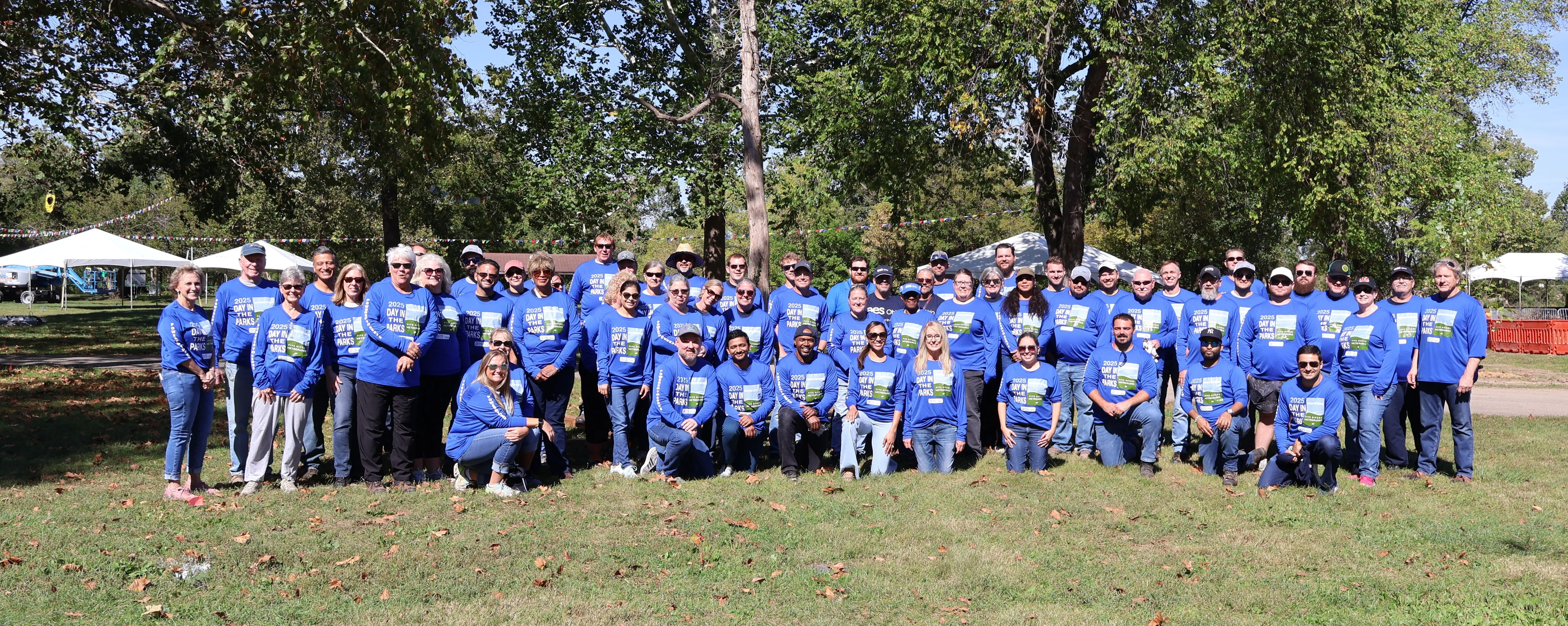 Large group of people wearing matching blue shirts with Day in the Parks printed on them, posing together outdoors in a park on a sunny day, with trees and event tents in the background.
