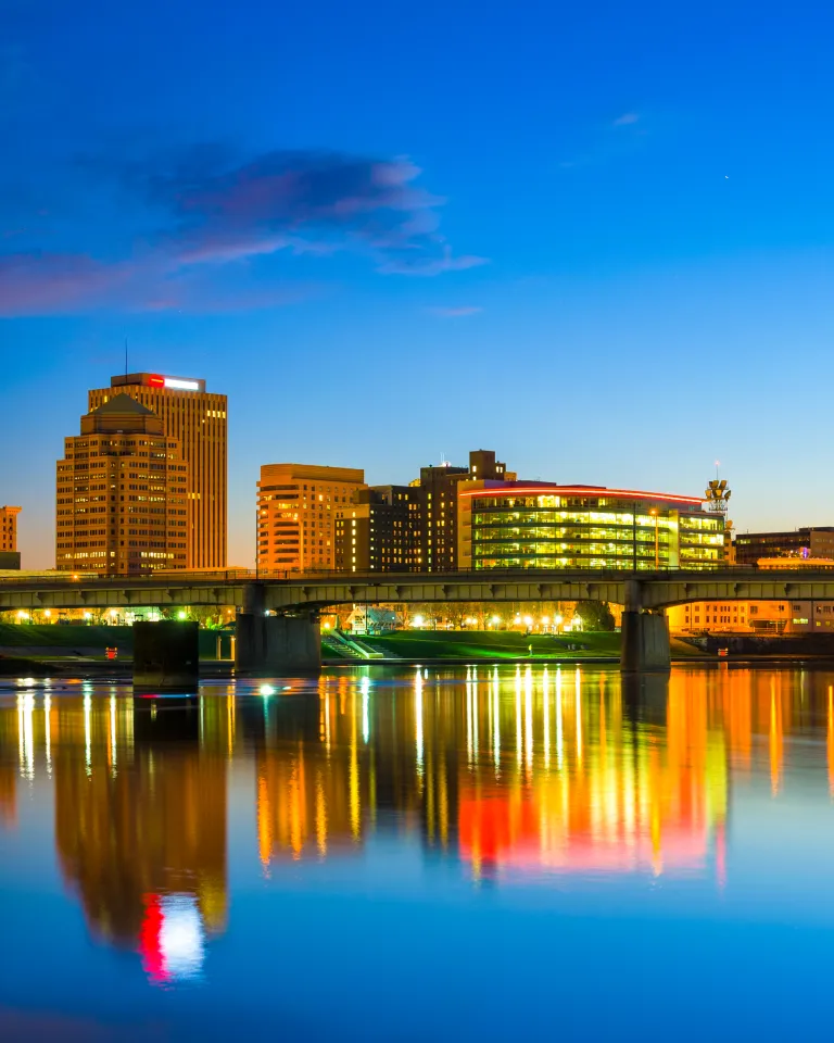 The skyline of Dayton, Ohio, is illuminated at night, reflecting vibrant city lights onto the calm waters of the river. A bridge spans the water, with blue-lit towers standing on either side. Modern and historic buildings rise in the background, their windows glowing against the deep blue sky. The scene is framed by the warm hues of the city lights, creating a striking contrast with the cool tones of the night.