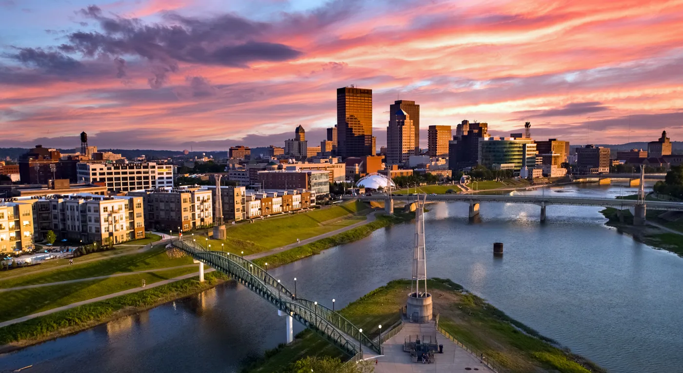 Aerial view of a Dayton, Ohio skyline at sunset with tall buildings, a river, pedestrian bridge, and colorful clouds in the sky.