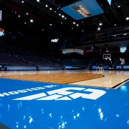 A basketball arena is set up for a March Madness event, with a polished wooden court reflecting bright overhead lights. The words "Hosted by AES" are printed on the blue sideline. A blue ladder stands near a Spalding basketball hoop. Players and staff are seen in the background, with one person taking a jump shot. The arena seating is mostly empty, and a large screen above the court displays the March Madness logo.