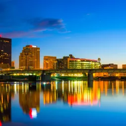 The skyline of Dayton, Ohio, is illuminated at night, reflecting vibrant city lights onto the calm waters of the river. A bridge spans the water, with blue-lit towers standing on either side. Modern and historic buildings rise in the background, their windows glowing against the deep blue sky. The scene is framed by the warm hues of the city lights, creating a striking contrast with the cool tones of the night.