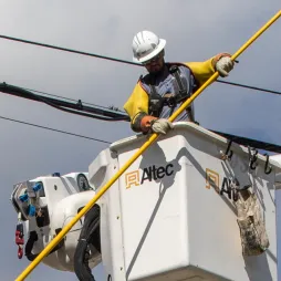 A utility worker wearing protective gear and a helmet uses an insulated pole to perform maintenance on overhead power lines. The worker is positioned in a white Altec bucket lift, elevated next to the cables, with a clear view of safety equipment and tools hanging from the lift. The sky is partly cloudy.