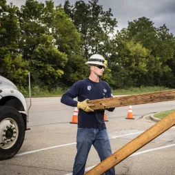 Utility worker in a hard hat and gloves carries a wooden crossarm from a service truck beside cable spools, with traffic cones marking a roadside work zone.