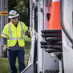 AES Ohio lineworker in high-visibility safety gear stands beside a utility truck with traffic cones, preparing for field operations on a sunny day.