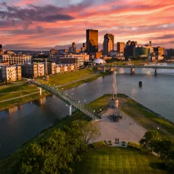 Aerial view of the Dayton, Ohio skyline at sunset with vibrant clouds, a river running through the city, modern buildings, a pedestrian bridge, and a green park area in the foreground.