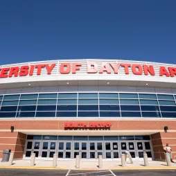 Front view of University of Dayton Arena with large red letters on the building and clear blue sky above. The entrance reads 'South Entry' and a few people are seen walking near the doors.
